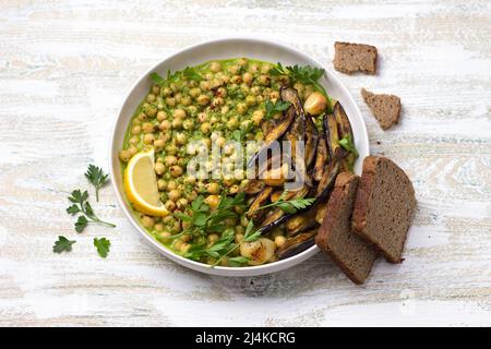 Grüne Kichererbsen mit gebackener Aubergine, Petersilie und Zitrone auf hellem Holztisch, rustikaler Stil, Draufsicht. Leckere gesunde hausgemachte Speisen Stockfoto