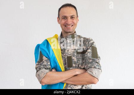 Ukrainischer Mann-Krieger, der mit einer Militär-Pixel-Uniform bekleidet ist, hält die gelb-blaue Flagge des Staates Ukraine und an der Kette ein kleines Wappen von Stockfoto