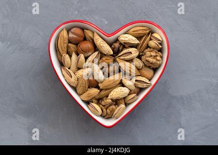 Verschiedene Nüsse in roter Schale Herz auf grauem Hintergrund geformt. Mandeln, Walnüsse, Haselnüsse, Pistazien. Gesunde Ernährung für das Herz. Draufsicht. Horizontal Stockfoto