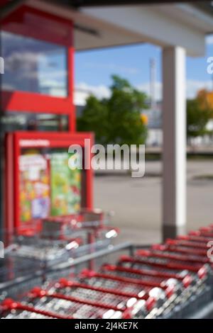 Nicht fokussiert. Rotes Werbeplakat und Reihe von Einkaufswagen im Eingangsbereich des Lebensmittelmarktes (Penny). Parkplatz im Hintergrund. Hoher pe-Wert Stockfoto