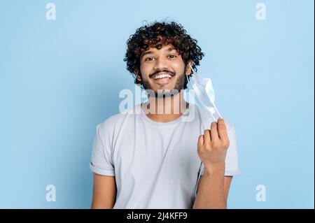Wiederherstellung, Abbruch der Quarantäne. Glücklicher indischer oder arabischer lockiger Kerl, zieht die schützende medizinische Maske aus, blickt freudig lächelnd auf die Kamera und steht auf einem isolierten blauen Hintergrund Stockfoto
