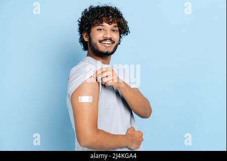 Gesundheitskonzept. Behandlung und Prävention, Impfkonzept. Zufriedener inder oder araber mit einer Bandhilfe auf der Schulter nach der Impfung gegen Covid 19, steht auf isoliertem blauem Hintergrund Stockfoto