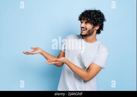 Hübscher attraktiver, lockiger inder- oder arabertyp im lässigen T-Shirt, sieht aus und zeigt die Hände zur Seite, auf leeren Kopierraum, steht auf isoliertem blauem Hintergrund und lächelt. Modellkonzept Stockfoto