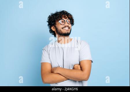 Hübscher, attraktiver indischer oder arabischer lockiger Kerl, trägt T-Shirt und Brille, steht auf einem isolierten blauen Hintergrund mit gekreuzten Armen, schaut weg und lächelt fröhlich Stockfoto