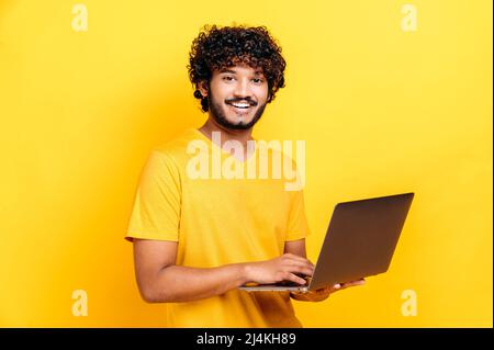 Ein attraktiver, zufriedener inder oder araber in T-Shirt, der einen offenen Laptop in der Hand hält, blickt freundlich auf die Kamera, während er vor einem isolierten orangefarbenen Hintergrund steht und fröhlich lächelt Stockfoto