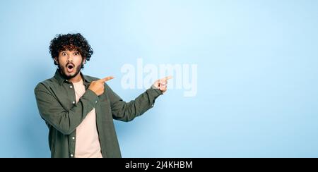 Panoramafoto von staunend schockiertem inder oder araber in legerer Kleidung, zeigt die Finger zur Seite auf einen nachgemockten leeren Raum, steht auf isoliertem blauem Hintergrund, schaut erstaunt auf die Kamera Stockfoto