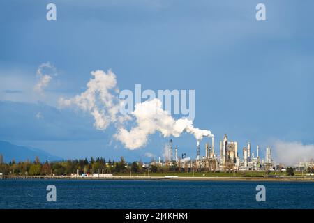 Dampf, der aus einem Schornstein in einer Erdölverarbeitungsanlage aufsteigt. Der weiße Dampf steht stark im Kontrast zu einem dunkelblauen Himmel über dem Industriekomplex. Stockfoto