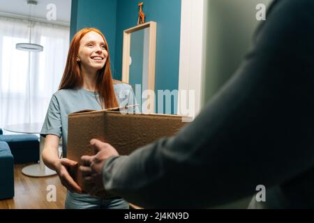 Lächelnd rothaarige junge Frau, die einen Karton von einem nicht erkennbaren Zusteller vor der Haustür der Wohnung annimmt Stockfoto