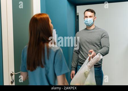 Kurier männlich in schützende Gesichtsmaske liefert Lebensmittelbeutel an weibliche Hausfrau Kunde auf Tür zu Hause. Stockfoto