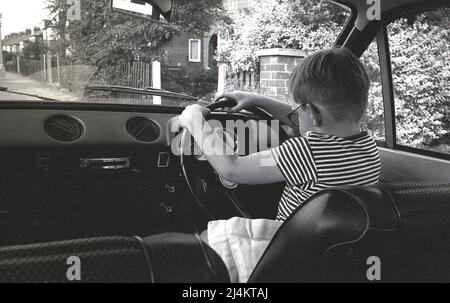 1970s, historisch, Blick von hinten, auf einen kleinen Jungen, der auf dem Fahrersitz eines Ford Escort Autos der damaligen Zeit sitzt, das Lenkrad hält und vorgibt zu fahren, England, Großbritannien. Stockfoto