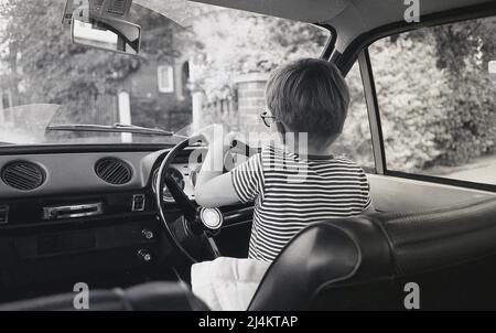 1970s, historisch, Blick von hinten, auf einen kleinen Jungen, der auf dem Fahrersitz eines Ford Escort Autos der damaligen Zeit sitzt, das Lenkrad hält und vorgibt zu fahren, England, Großbritannien. Stockfoto