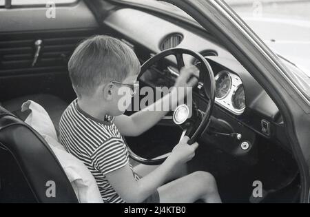 1970, historisch, Seitenansicht, Nahaufnahme eines Jungen, der auf dem Fahrersitz eines Ford Escort Autos der damaligen Zeit sitzt, das Lenkrad hält und vorgibt zu fahren, England, Großbritannien. Stockfoto