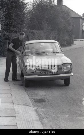 1970, historisch, ein kleiner Junge auf dem Fahrersitz eines Autos der Ära, ein Ford Escort, der das Lenkrad hält, vorgibt zu fahren, Vater vor dem Auto, das auf einer ruhigen Vorstadtstraße, England, Großbritannien, geparkt ist. Stockfoto