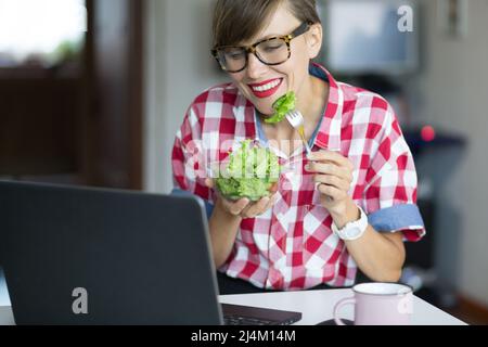 Schöne kaukasische Frau, die zu Hause Salat isst, Pause zwischen der Arbeit Stockfoto