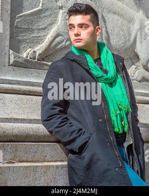 In einer schwarzen Kapuzenjacke, einem grünen Schal, einer blauen Hose und langen Lederstiefeln sitzt ein junger, gutaussehender Kerl da und entspannt sich. Stockfoto