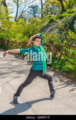 In einem grünen Langarmshirt, Schal, schwarzen Hosen und Lederstiefeln, die Arme strecken und die Beine umdrehen, trainiert ein junger Mann outsid Stockfoto