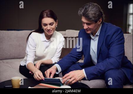 Entspanntes Paar mittleren Alters, das Farbstoffmuster für die Polsterung der Möbel pflückt und auf dem Sofa im Ausstellungsraum des Möbelhauses sitzt Stockfoto