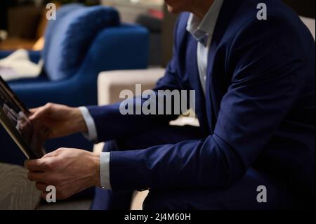 Zugeschnittenes Bild. Hände eines Mannes im Geschäftsanzug, der einen Katalog hält und auf einem Sofa im Showroom eines Polstermöbelgeschäftes sitzt. Klein, Bussin Stockfoto