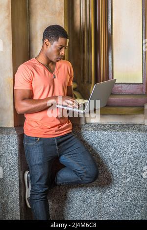 Ein junger, hübscher schwarzer Student, der sich in einem hellorangenen Kurzarm-T-Shirt mit V-Ausschnitt und Jeans kleidet, steht an einem kleinen Fenster und arbeitet an einem Laptop Stockfoto