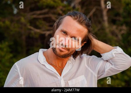 Porträt eines gutaussehenden brutalen Mannes mit langen Haaren bei Sonnenuntergang, die blauen Augen, eine leichte Borste, er ist in einem weißen Hemd mit einem kurzen Ärmel, grün gekleidet Stockfoto