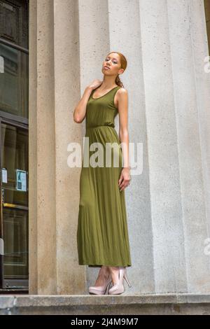 In einem grünen Maxi Tank Dress, cremigen High Heels, überkreuzten Beinen und einer Hand, die ihre Schulter berührt, steht ein junges, schwarzes Mädchen zur Seite Stockfoto