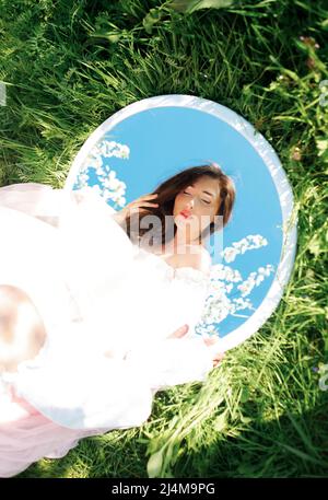 Junge schöne Frau, Spiegelung im Spiegel. Kreatives Frühlingsfoto im blühenden Garten. Stockfoto