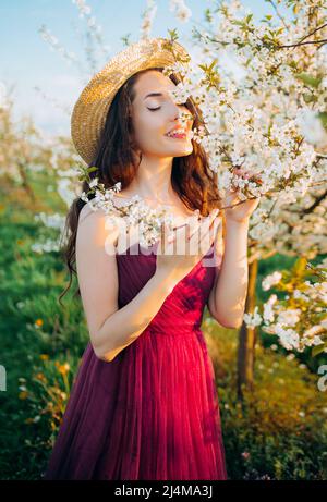 Porträt eines verträumten Mädchens im Frühlingspark unter den blühenden weißen Bäumen bei Sonnenuntergang. Frau in Strohhut und purpurfarbenem Kleid, Kirschblüten. Feder s Stockfoto