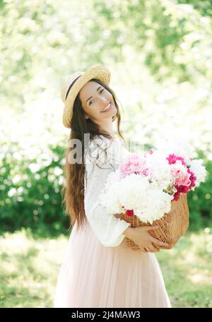 Fröhliches lächelndes Mädchen in einem Strohhut, das einen Korbkorb mit weißen und rosa Pfingstrosen-Blumen in den Straßen einer europäischen Stadt hält. Lifestyle Stockfoto