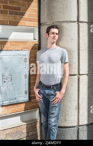 In einem grauen T-Shirt und einer blauen Jeans kleidet, steht ein junger, gutaussehender Kerl in einer Ecke der Straße, mit den Augen zuckend und tief denkend. Stockfoto