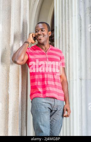 Ein junger schwarzer Mann, der sich in einem roten, pinken Streifen mit Henley V Neck T-Shirt, einer grauen Hose und einem Armband kleidet, steht an einer Säule, lächelt und macht ein P Stockfoto