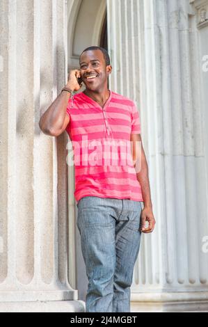 Ein junger schwarzer Mann, der sich in einem roten, pinken Streifen mit Henley V Neck T-Shirt, einer grauen Hose und einem Armband kleidet, steht an einer Säule, lächelt und redet weiter Stockfoto