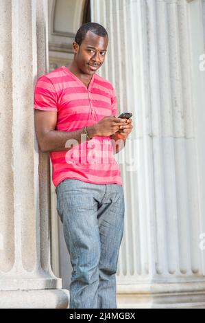 Ein junger schwarzer Mann, der sich in einem roten, pinken Streifen mit Henley V Neck T-Shirt, einer grauen Hose und einem Armband kleidet, steht an einer Säule und überprüft Nachrichten auf h Stockfoto