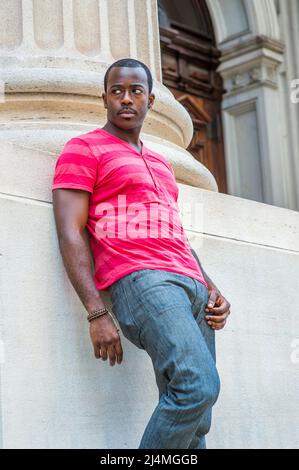 In rot, rosa Streifen Henley V Neck T-Shirt, graue Hose, trägt ein Armband, ein junger schwarzer Kerl steht gegen die Basis einer Säule outsi Stockfoto