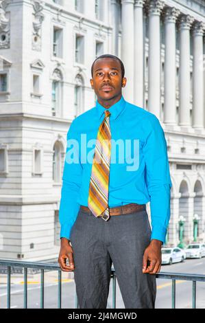 Ein junger, schwarzer Geschäftsmann, der sich mit einem hellblauen Hemd, einer grauen Hose und einer bunten Krawatte ankleidet, steht vor einem Bürogebäude und freut sich nach vorne Stockfoto