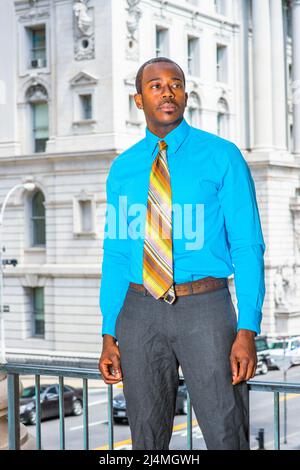 Ein junger, schwarzer Geschäftsmann, der sich mit einem hellblauen Hemd, einer grauen Hose und einer bunten Krawatte ankleidet, steht vor einem Bürogebäude und freut sich nach vorne Stockfoto