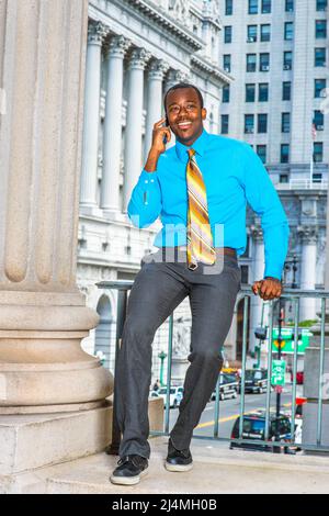 Ein junger, schwarzer Geschäftsmann sitzt vor einem Bürogebäude und ruft mit einem hellblauen Hemd, einer grauen Hose, Lederschuhen und einer Krawatte an Stockfoto