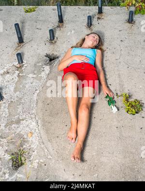 Ein hübsches blondes Mädchen, das sich in hellblauem Tank-Top, roten Shorts und barfuß kleidet, eine Hand mit einer weißen Rose, liegt auf einer alten Baustelle mit Witz Stockfoto