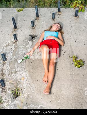 Ein hübsches blondes Mädchen, das sich in hellblauem Tank-Top, roten Shorts und barfuß kleidet, eine Hand mit einer weißen Rose, liegt auf einer alten Baustelle mit Witz Stockfoto