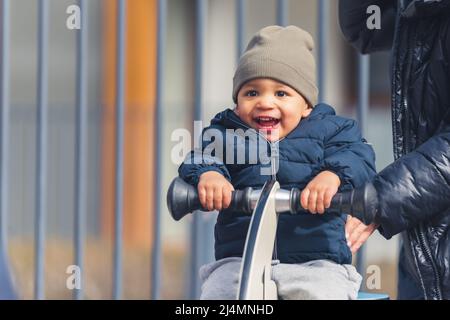 Outdoor-Porträt eines extrem glücklich Smiley jungen Biracial Kleinkind in einem grauen Hut und warmen Mantel sitzen auf einem Spielplatz Frühling Reiter und Spaß haben. Hochwertige Fotos Stockfoto