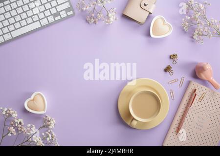 Gemütlicher femininer Schreibtisch mit Tastatur, Kaffeetasse, herzförmige Kerzen, rosa Notizbuch, Büromaterial, Frühlingsblumen auf pastellviolettem Hintergrund Stockfoto