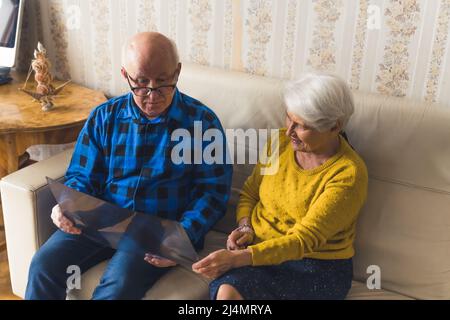 Die kaukasischen Ehegatten im Ruhestand betrachten die Röntgenergebnisse, während sie auf einem beigefarbenen Ledersofa sitzen. Unterstützung während der Krankheit. Hochwertige Fotos Stockfoto