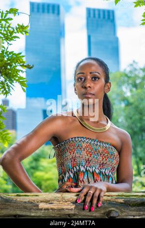 In einem trägerlosen, farbenfrohen Kleid mit goldenem Halsring steht eine hübsche schwarze Frau in einem kleinen Wald in einer Großstadt und wartet auf dich Stockfoto