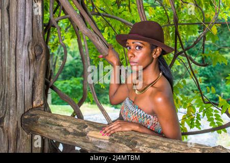 Mit einem braunen Outback-Hut, einem goldenen Halsring, steht eine hübsche schwarze Frau draußen in einem kleinen Wald und schaut dich an. Stockfoto