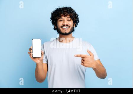 Gutaussehender, glücklicher inder oder araber, schaut auf die Kamera, hält das Smartphone mit dem weißen, leeren Bildschirm zur Präsentation in der Hand und zeigt mit dem Finger darauf, steht auf einem isolierten blauen Hintergrund und lächelt Stockfoto