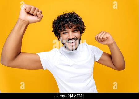 Fröhlicher, positiver inder oder araber mit lockigen Haaren, trägt ein weißes T-Shirt, blickt fröhlich auf die Kamera, spreizt seine Arme zur Seite, steht über einem isolierten orangefarbenen Hintergrund, schaut auf die Kamera, lächelt Stockfoto