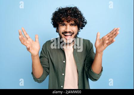 Entmutigter indischer oder arabischer verwirrter, lockiger junger Mann, der emotional mit seinen Händen gestikulierte, neugierig auf die Kamera schaute und auf einem isolierten blauen Hintergrund stand Stockfoto