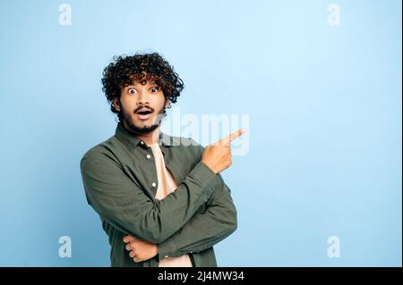 Überrascht schockierter inder oder araber in legerer Kleidung, erstaunt blickt auf die Kamera und zeigt den Finger zur Seite auf den Raum zur Präsentation, steht auf isoliertem blauem Hintergrund, lächeln. Konzept für den Kopierraum Stockfoto