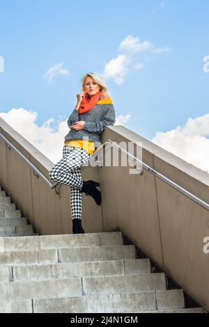 Ein junges blondes Mädchen trägt eine gelbe Unterwäsche, einen grauen modischen Pullover, eine Hose mit schwarz-weißem Muster, einen roten Schal, eine Sonnenbrille in der Hand Stockfoto