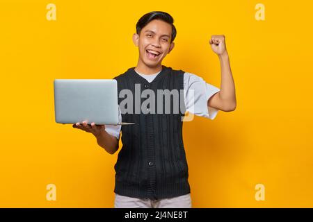 Aufgeregt asiatischen Mann hält Laptop und feiert Sieg lächelnd über gelben Hintergrund Stockfoto
