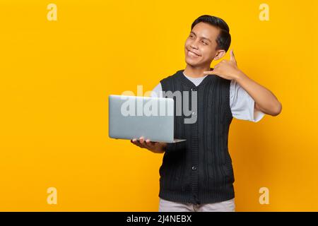 Porträt einer lächelnden jungen asiatischen Frau, die ihren Laptop hält und mit dem Finger eine Telefongeste macht Stockfoto